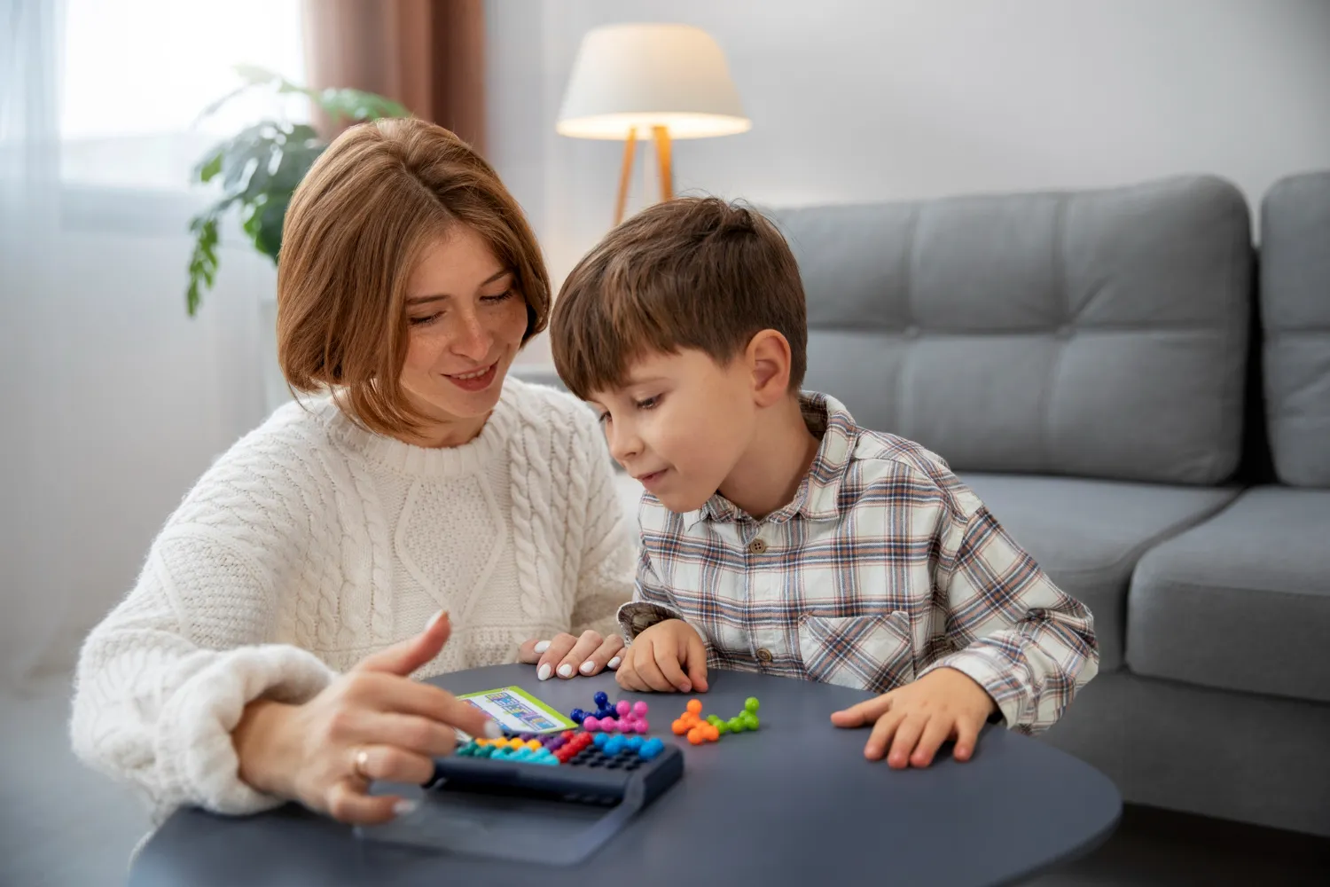 Madre e hijo interactuando con un juego educativo en una mesa, rodeados de colores y juguetes, en un ambiente acogedor y luminoso.