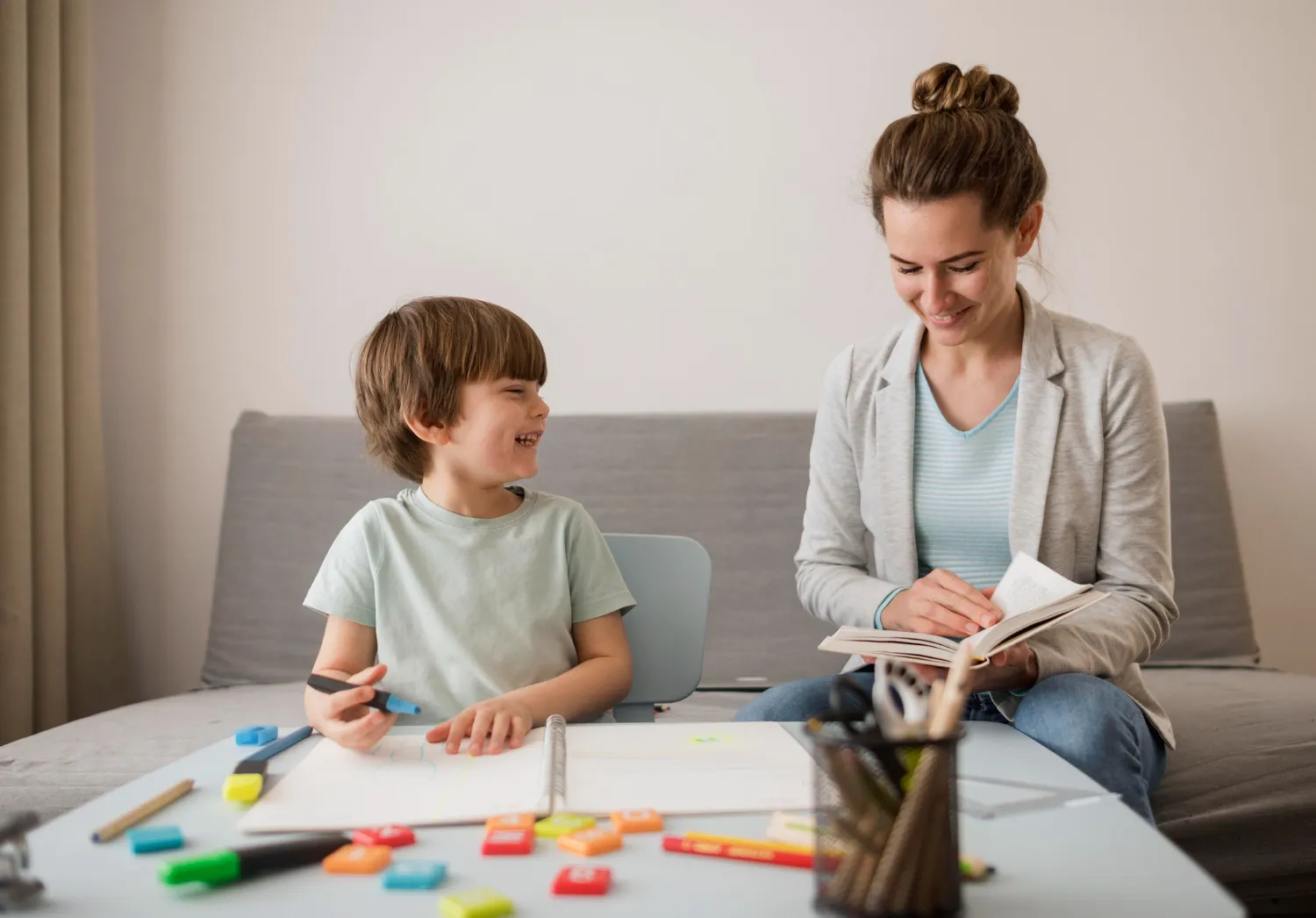 Mujer y niño sonrientes en una mesa de estudio, rodeados de materiales de arte y libros. El niño sostiene un marcador y dibuja, mientras la mujer lee. Ideal para ilustrar actividades educativas y de creatividad en familia.