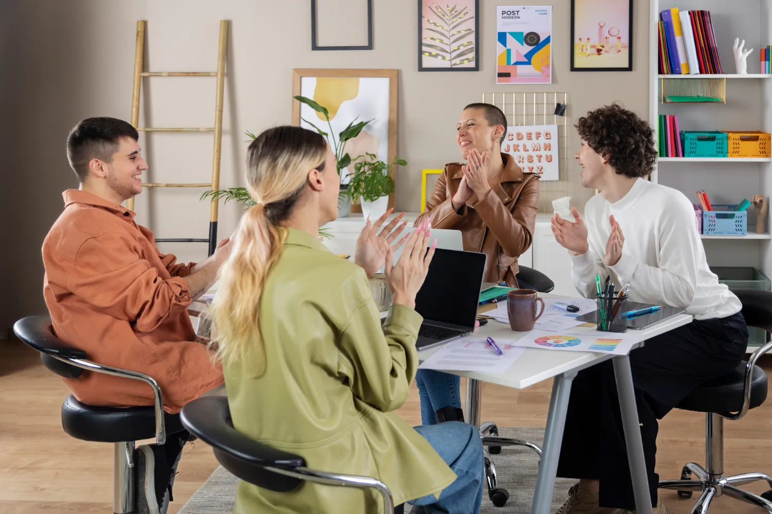Grupo de cuatro personas aplaudiendo en una reunión creativa, sentados alrededor de una mesa con documentos, un laptop y materiales de oficina en un ambiente moderno y luminoso.
